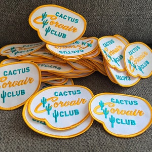 May include: A collection of embroidered patches with the text "CACTUS CORVAIR CLUB". The patches are white with a yellow border and feature a turquoise cactus and lettering. The patches are scattered on a gray surface.