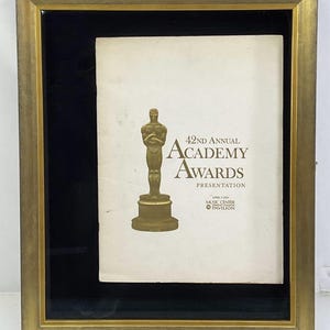 May include: Framed presentation from the 42nd Annual Academy Awards. The gold-toned frame surrounds a black matting, highlighting the printed program. The program features the Oscar statuette and text that reads "42nd Annual Academy Awards Presentation."