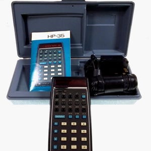 May include: A black HP-35 scientific calculator with a blue display and silver buttons. The calculator is in a gray carrying case with a power adapter and instruction manual.