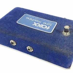 May include: A blue FOXX Tone Machine guitar effects pedal. The pedal has a blue felt-like exterior with a silver label that reads "FOXX Tone Machine". It features input and output jacks, a footswitch, and control knobs. The pedal is designed for guitar effects.