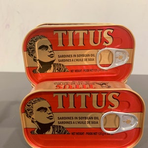 May include: Two red and gold cans of Titus sardines in soybean oil. Each can has a picture of a man with a crown on his head. The text on the can reads "Sardines in Soybean Oil" and "Sardines a l'huile de soja". The net weight of each can is 125 grams (4.4 ounces).