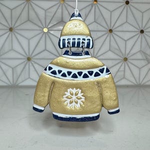 May include: A gold-colored sweater ornament with a matching hat, both trimmed with navy blue and white accents. The sweater features a white snowflake design on the front. The ornament is hanging by a white string.