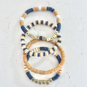 Puede incluir: Pila de cuatro pulseras de cuentas. Las pulseras presentan una combinación de cuentas azul marino, blancas y beige. Algunas pulseras deletrean "KENT STATE" y "EAGLES" con cuentas de letras. Las pulseras están apiladas sobre una superficie blanca.