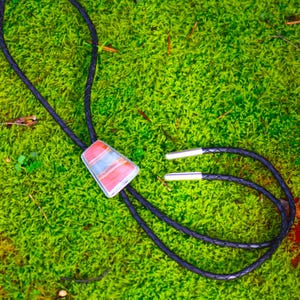 May include: A bolo tie with a rectangular, multi-colored stone pendant in a silver setting. The stone features shades of pink, blue, and white. The black braided leather cord has silver tips and a sliding clasp.