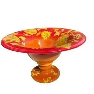 May include: A decorative, pedestal bowl with a wide, shallow rim. The bowl is painted with a gradient of orange to red, and adorned with yellow floral designs. The base and stem are a matching orange color.