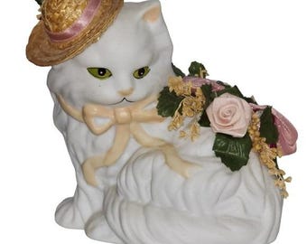 Puede incluir: Figura de gato de cerámica blanca con ojos verdes, que lleva un sombrero de paja con una cinta rosa y un lazo crema. El gato está adornado con rosas rosas y vegetación, creando una pieza decorativa caprichosa.