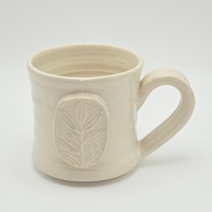 May include: A cream-colored ceramic mug with a glossy finish and a wide, curved handle. The exterior features a raised, oval-shaped medallion on the front, embossed with a detailed botanical design of leaves and stems against a textured background.