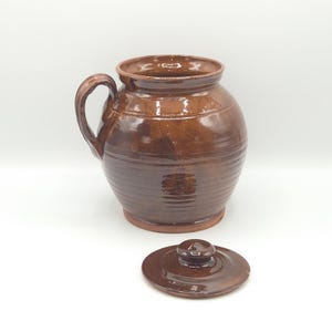 May include: A brown glazed ceramic jar with a single side handle and a matching circular lid. The jar features a rounded body with horizontal ribbed detailing along the lower section and a smooth, glossy finish throughout the exterior surface.