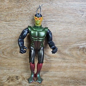 May include: A humanoid action figure with a yellow-green head, orange eyes, and a dark gray antenna. The figure features a metallic green chest plate, black arms, dark gray upper legs, and maroon lower legs with pointed, claw-like feet.