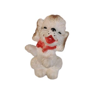 Peut inclure: Une figurine de caniche blanc avec des oreilles et une touffe de poils teintées de brun, ornée d'un nœud rouge vif autour du cou. Le chien est représenté debout avec une patte avant levée, des yeux noirs, un nez noir et une gueule rouge ouverte.