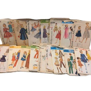 May include: A collection of vintage sewing pattern envelopes featuring various illustrations of clothing designs. The envelopes are primarily white with printed text including brand names like Simplicity and Butterick, along with pattern numbers and prices.