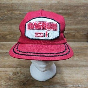 May include: A red baseball cap featuring a white patch on the front with the red embroidered text MAGNUM and the Case IH logo below it. The cap has a flat brim with two rows of black stitching along the edge and a structured crown design.