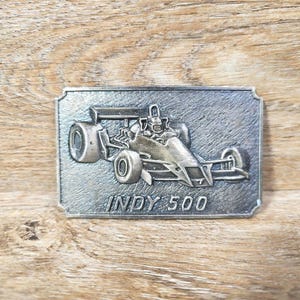 May include: A rectangular silver-toned metal belt buckle featuring a raised relief design of an open-wheel race car. The text INDY 500 is embossed in capital letters below the vehicle. The surface has a brushed, textured finish throughout.