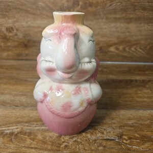 May include: A ceramic elephant-shaped vase featuring a cream-colored head with pink accents, closed eyes, and a trunk. The body is pink with a floral-patterned bib detail. The top opening is circular, and the overall finish has a glossy glaze.