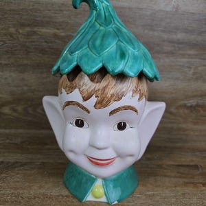 May include: A ceramic cookie jar shaped like an elf head with a pointed green leaf-patterned hat lid. The elf has light skin, brown hair, large pointed ears, and a green collar with a single yellow button. The facial features are painted on.