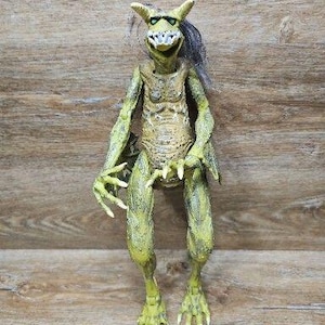 May include: A green, textured creature action figure with pointed ears, a wide mouth displaying sharp white teeth, and long, clawed fingers. The figure has a hunched posture, a lean torso, and wispy dark hair extending from the back of its head.