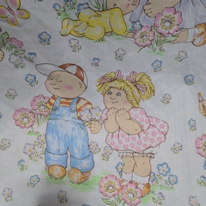 May include: A white fabric featuring a repeating pattern of cartoon children with round faces and rosy cheeks. The characters are depicted in colorful outfits, including blue overalls and pink dresses, surrounded by illustrations of pink and blue flowers.