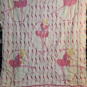 May include: A white quilted bedspread featuring a repeating pattern of blonde ballerinas in pink tutus and bodices. The design includes vertical wavy pink ribbons and small pink rose accents throughout, finished with a decorative rose border.