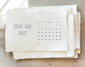May include: A stack of off-white, textured paper cards with deckled edges. The top card features the text JANUARY 2027 in a serif font, followed by a grid of the month's dates starting with the letter S for Sunday through S for Saturday.