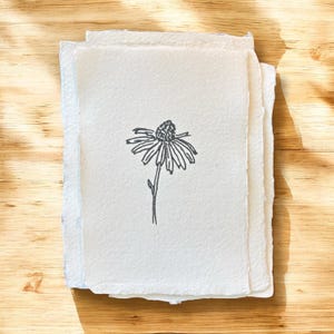May include: A stack of rectangular, off-white paper sheets with rough, deckled edges. The top sheet features a centered, black ink line drawing of a single coneflower with a textured, rounded center and elongated, drooping petals on a thin stem.