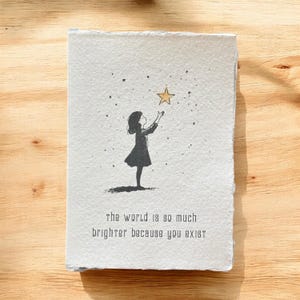 May include: A black and white illustration on textured paper. It depicts a silhouette of a person reaching for a gold star. The text at the bottom reads: "the world is so much brighter because you exist."