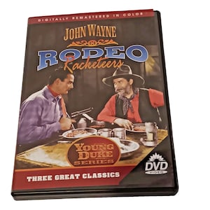 May include: DVD case for "Rodeo Racketeeers" starring John Wayne. The cover features a scene of two men at a table, with the text "Young Duke Series" and "Three Great Classics". The DVD case is black.