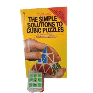 Può includere: Un libro giallo intitolato "The Simple Solutions to Cubic Puzzles" con una mano che tiene un puzzle piramidale colorato. Un piccolo puzzle a cubo avvolto con un design Angry Birds è di fronte al libro.