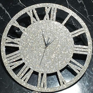 Puede incluir: Un reloj de pared circular cubierto de pequeños cristales plateados reflectantes. La esfera presenta grandes números romanos y dos manecillas finas de color plateado. Toda la superficie, incluyendo el marco y los números, está incrustada con piedras brillantes.