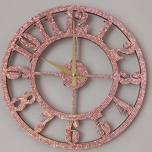 Puede incluir: Un reloj de pared redondo y decorativo con una esfera cubierta de pedrería rosa y un marco marrón oscuro. El reloj presenta manecillas doradas y números romanos. El diámetro del reloj es de aproximadamente 30 cm.