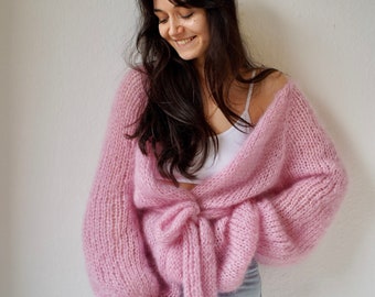 May include: A pink knitted cardigan with a tie at the waist. The cardigan has long sleeves and a relaxed fit.