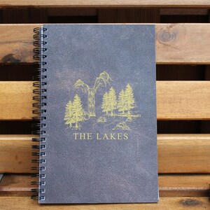 May include: A dark gray spiral bound notebook with a gold illustration of trees and the text "THE LAKES" on the front cover.