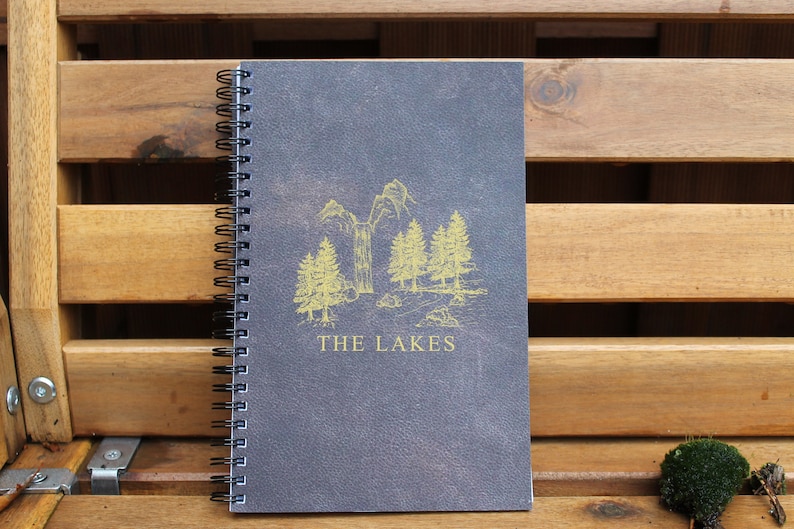 May include: A dark gray spiral bound notebook with a gold illustration of trees and the text "THE LAKES" on the front cover.