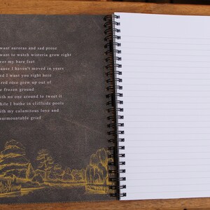 May include: A black and gray spiral bound notebook with a dark gray cover featuring a poem about auroras, wisteria, and grief. The cover also has a gold-colored illustration of a river and trees.
