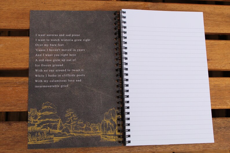 May include: A black and gray spiral bound notebook with a dark gray cover featuring a poem about auroras, wisteria, and grief. The cover also has a gold-colored illustration of a river and trees.