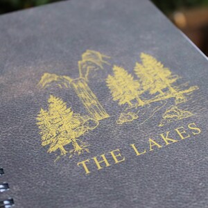 May include: A dark gray spiral bound notebook with a gold illustration of trees and the text "THE LAKES" on the cover.