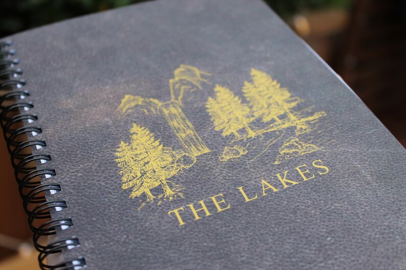 May include: A dark gray spiral bound notebook with a gold illustration of trees and the text "THE LAKES" on the cover.