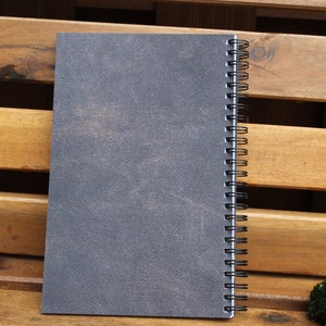 May include: A gray spiral bound notebook with a textured cover. The notebook is sitting on a wooden surface.
