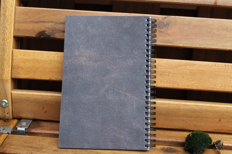 May include: A gray spiral bound notebook with a textured cover. The notebook is sitting on a wooden surface.