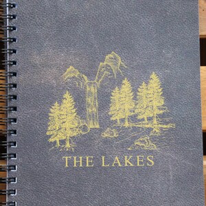 May include: A gray spiral bound notebook with a yellow illustration of a mountain scene and the text "THE LAKES" on the front cover.