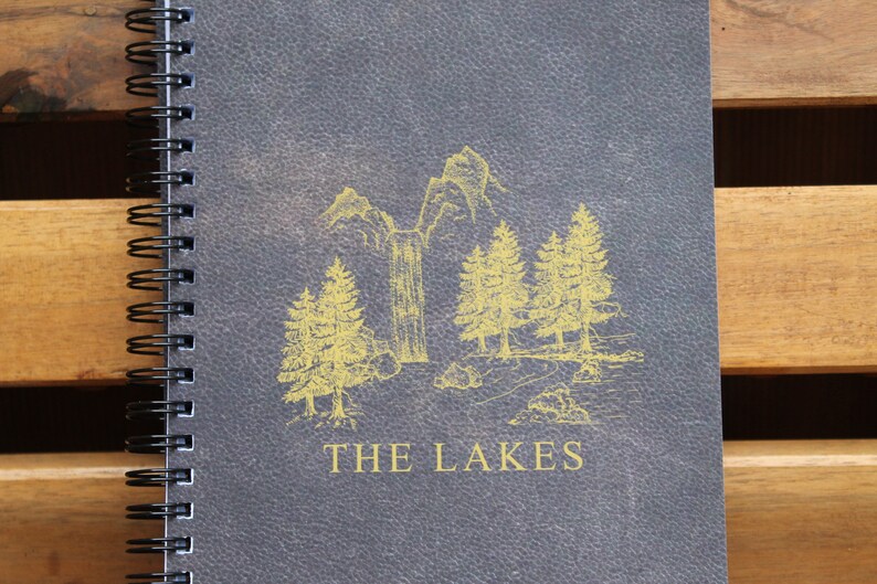 May include: A gray spiral bound notebook with a yellow illustration of a mountain scene and the text "THE LAKES" on the front cover.