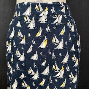 Può includere: Gonna a tubino blu navy con un motivo ripetuto di barche a vela bianche e gialle. La gonna ha una vita alta e una silhouette aderente. Le barche a vela sono raffigurate con vele bianche e dettagli gialli, creando un tema nautico.