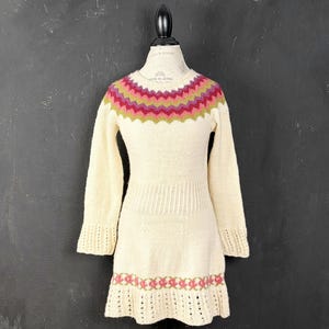 Peut inclure: Une robe en tricot crème à manches longues avec un col rond orné d'un motif en zigzag magenta et vert olive. La robe présente une taille côtelée, des manches longues à poignets côtelés et un ourlet agrémenté d'un motif floral assorti.