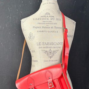 May include: A bright red satchel bag with a shoulder strap, displayed on a mannequin. The bag has a rectangular shape with a flap closure and two silver buckles. The strap is also red, and the bag appears to be made of leather.