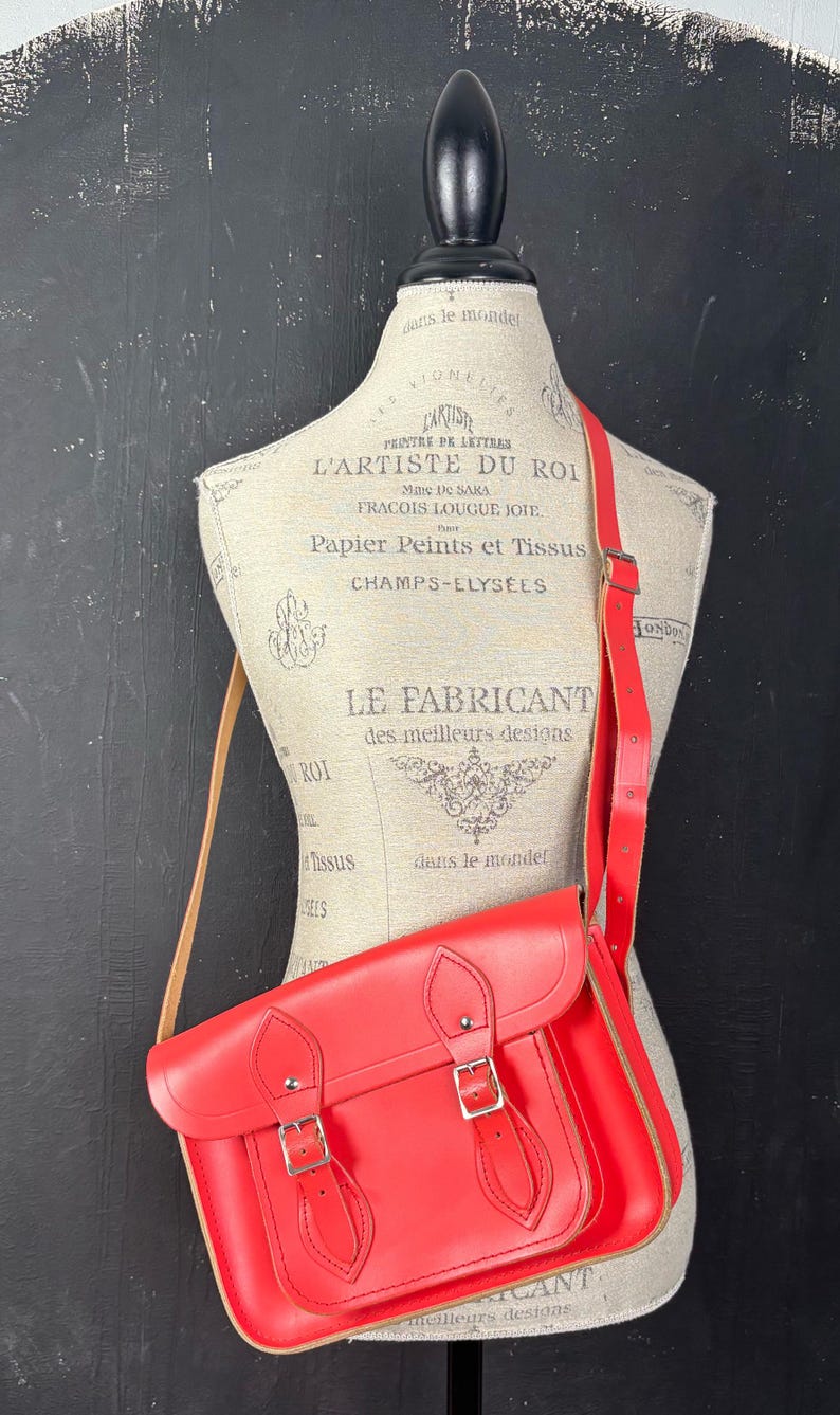 May include: A bright red satchel bag with a shoulder strap, displayed on a mannequin. The bag has a rectangular shape with a flap closure and two silver buckles. The strap is also red, and the bag appears to be made of leather.