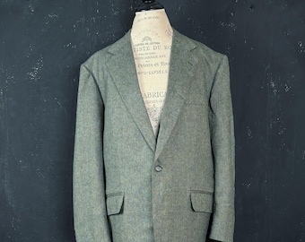 Pode incluir: Um blazer verde-sálvia com acabamento texturizado, apresentando lapela entalhada, fecho frontal de um botão e dois bolsos com aba na cintura. A peça é exibida em um torso de manequim bege com detalhes de texto impressos.