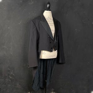 Könnte beinhalten: Ein schwarzer Frack auf einer Schaufensterpuppe. Die formelle Jacke hat ein fallendes Revers, einen zweireihigen Knopfverschluss und lange Schöße. Die Schaufensterpuppe steht vor einem dunklen Hintergrund.