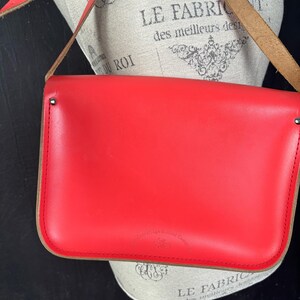 May include: A bright red leather satchel bag with a long shoulder strap. The bag has a rectangular shape with rounded corners and a contrasting tan trim. The strap is also red and appears to be adjustable. The bag is displayed on a mannequin.
