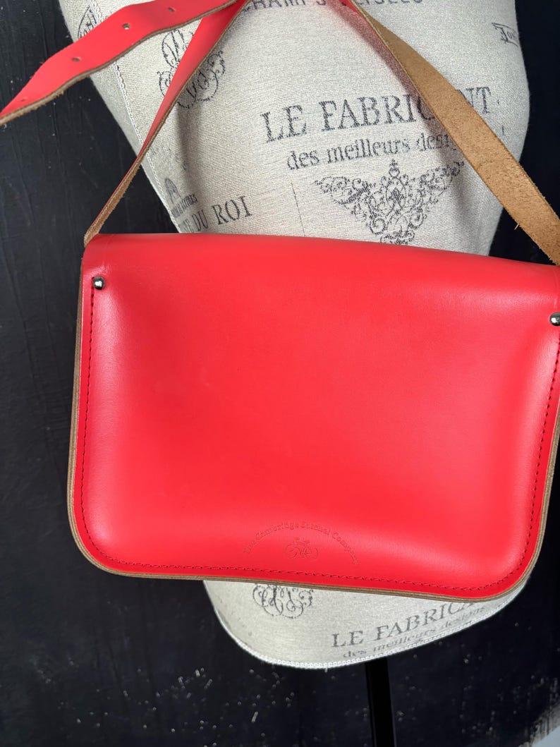May include: A bright red leather satchel bag with a long shoulder strap. The bag has a rectangular shape with rounded corners and a contrasting tan trim. The strap is also red and appears to be adjustable. The bag is displayed on a mannequin.