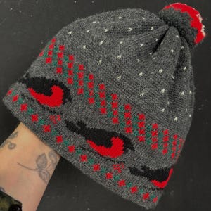 May include: A charcoal gray knit beanie featuring a red and green bird pattern around the base, a small white speckled design on the upper section, and a red pom-pom on top. The hat is constructed from a textured wool-like material.