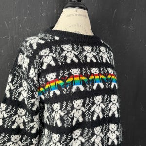 May include: Black knitted sweater featuring a repeating pattern of white teddy bears and snowflakes. A rainbow band of teddy bears is across the chest. The sweater has a crew neck and long sleeves.